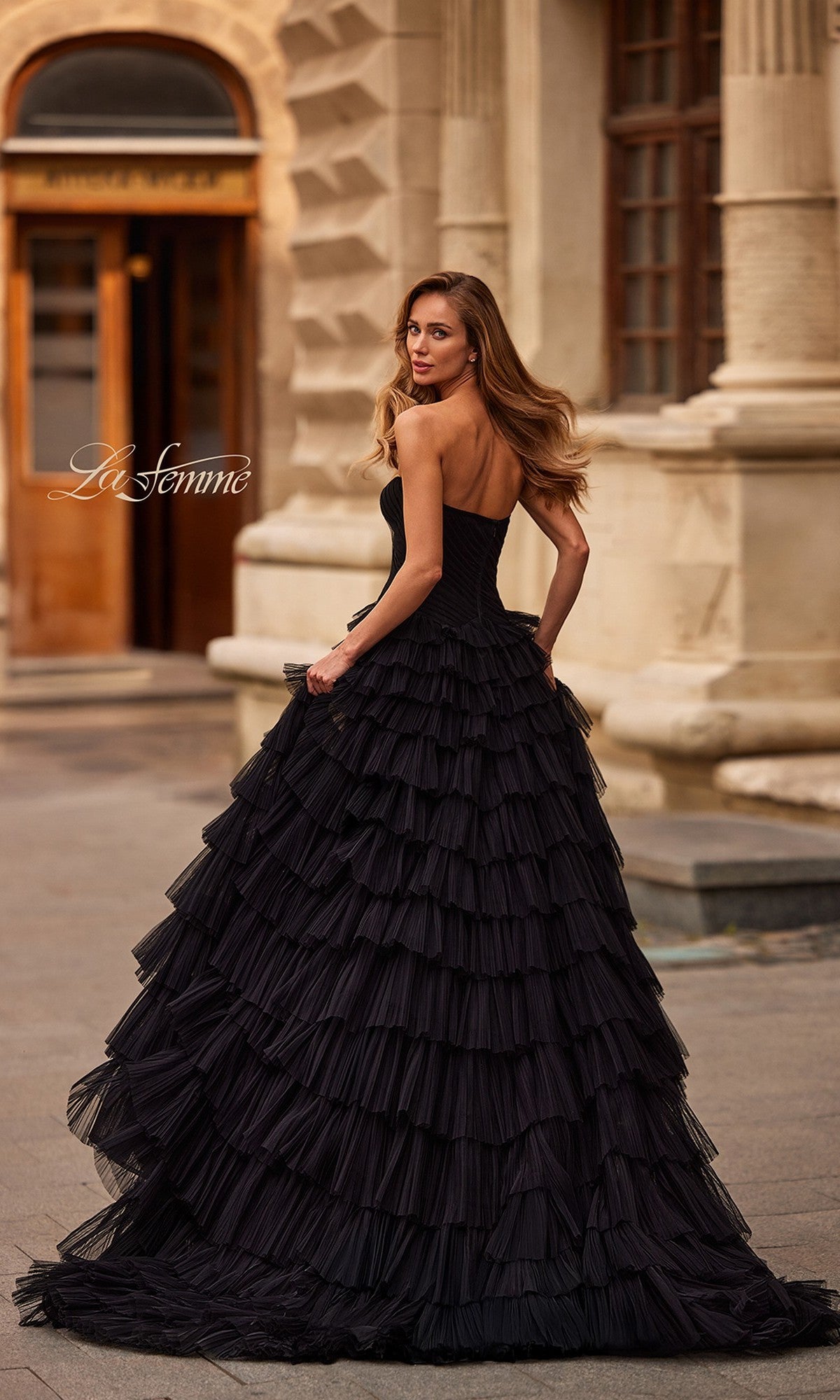 A woman wearing the La Femme 33202 long black strapless prom dress with a pleated bodice and ruffled skirt stands outdoors before an ornate building, glancing over her shoulder.